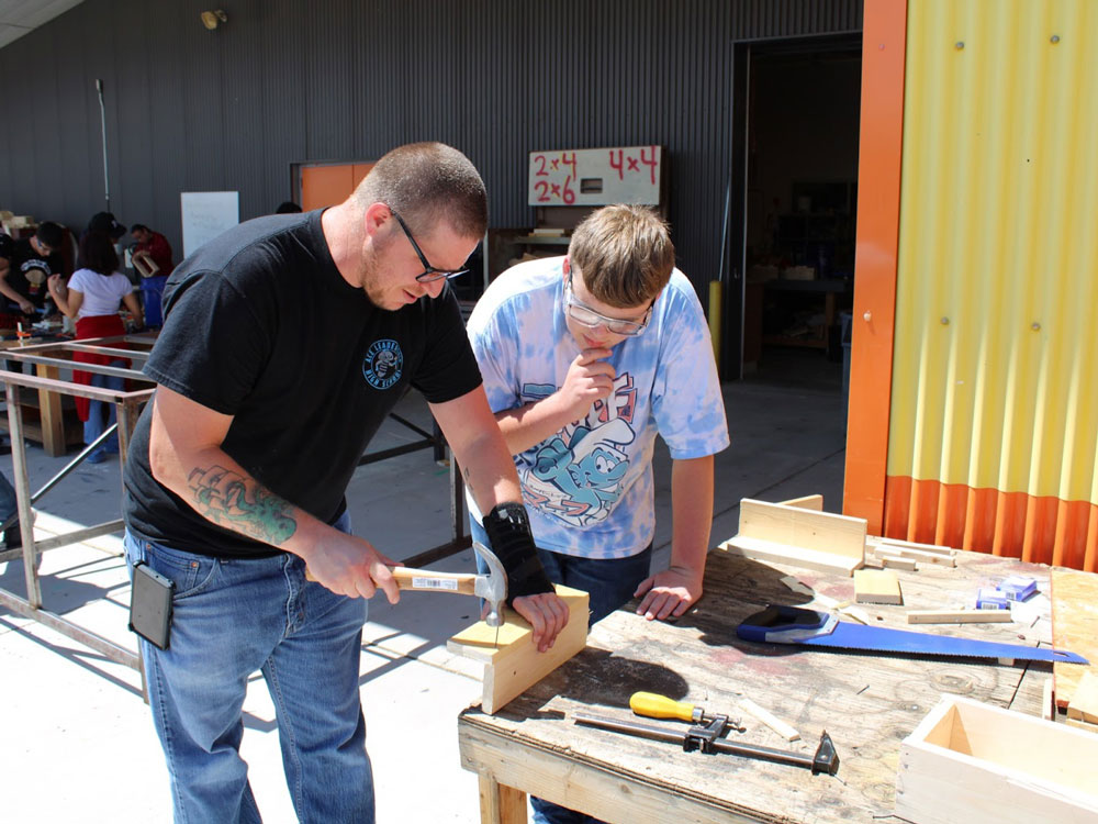 Decorative photo of an educator hammering a nail into timber with a student looking on.